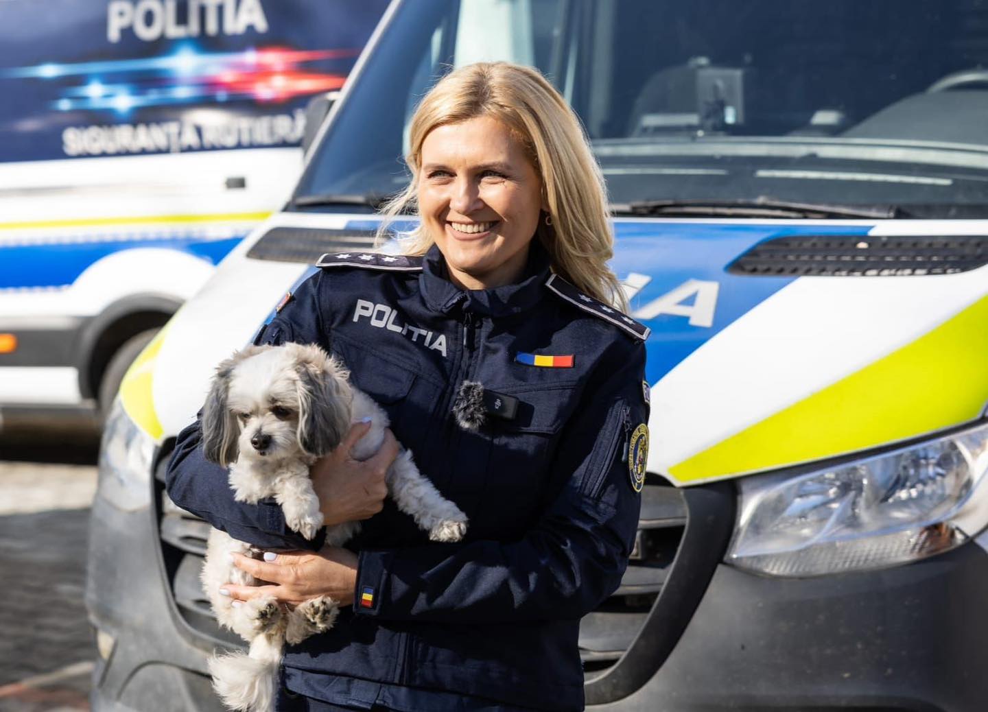 O polițistă ține în brațe un câine, iar pe fundal sunt mașini de Poliție.
