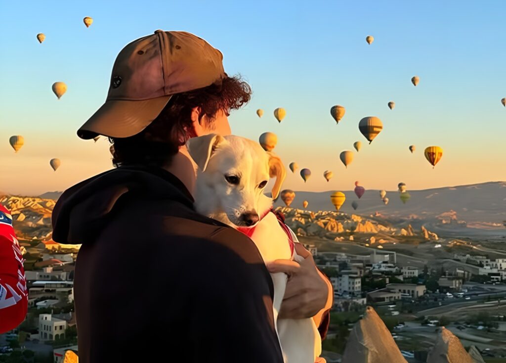 Un tânăr își ține câinele în brațe și se uită în zare la baloane cu aer cald