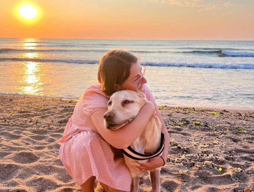 Un câine este ținut în brațe de stăpâna lui, pe plajă, la apus