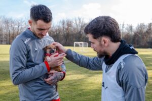 Doi jucători ai echipei de fotbal Dinamo București mângâie un cățel